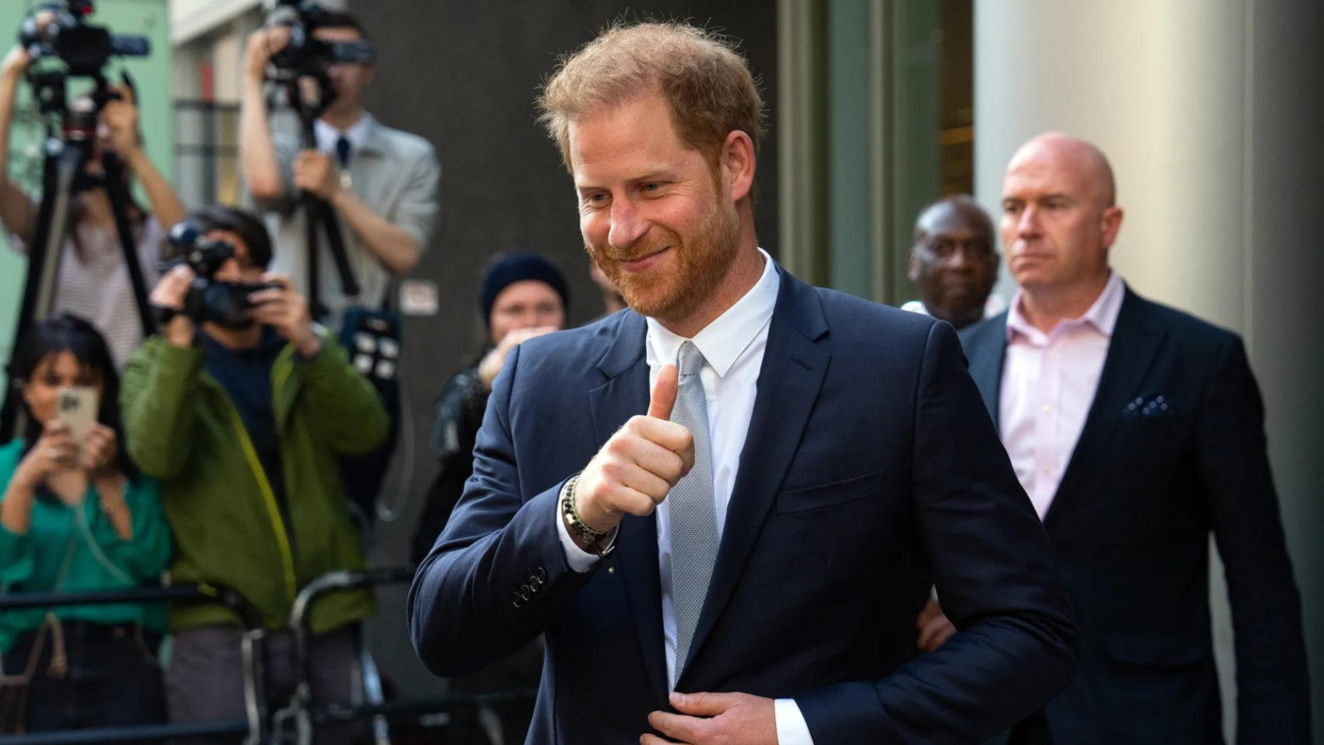 Prince Harry in a suit with a slight smile making a fist pump gesture as photographers capture the moment.