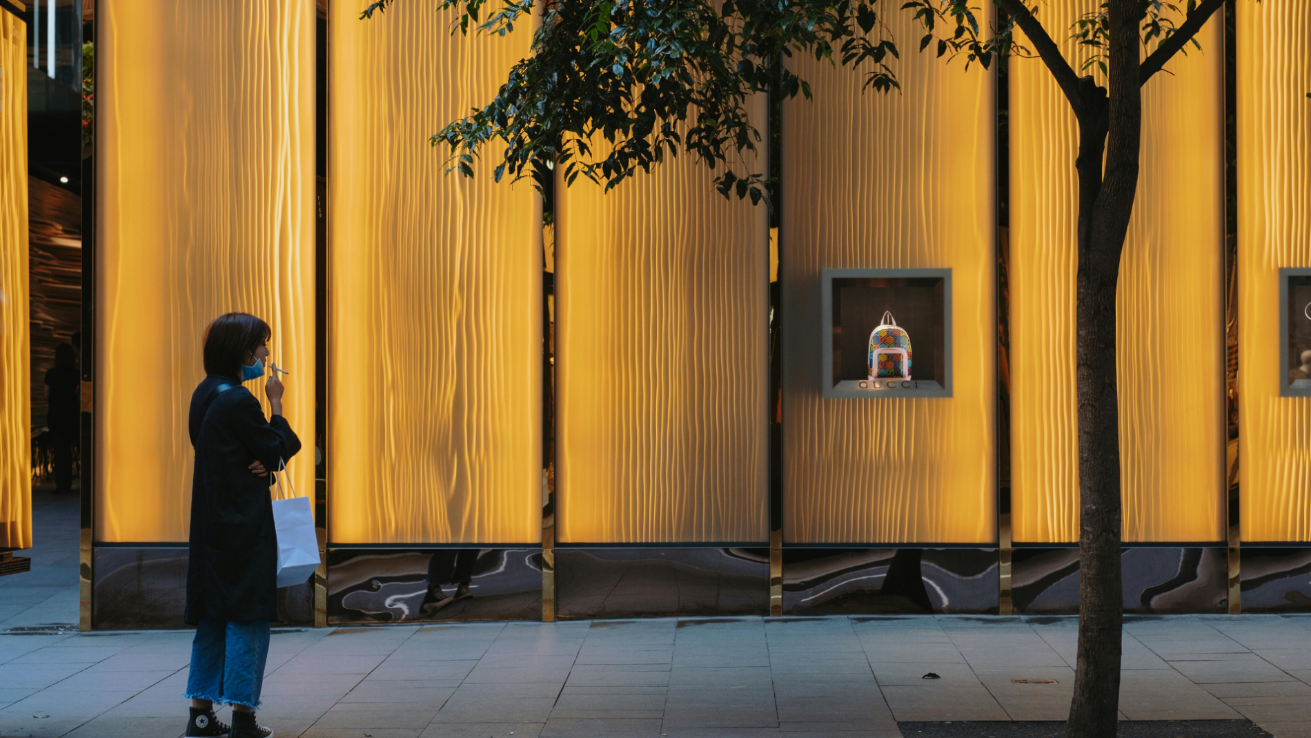 A woman in a dark coat stands on a sidewalk, contemplatively looking at a handbag in a warmly-lit luxury store window display, with a lone tree adding a touch of greenery to the urban scene.