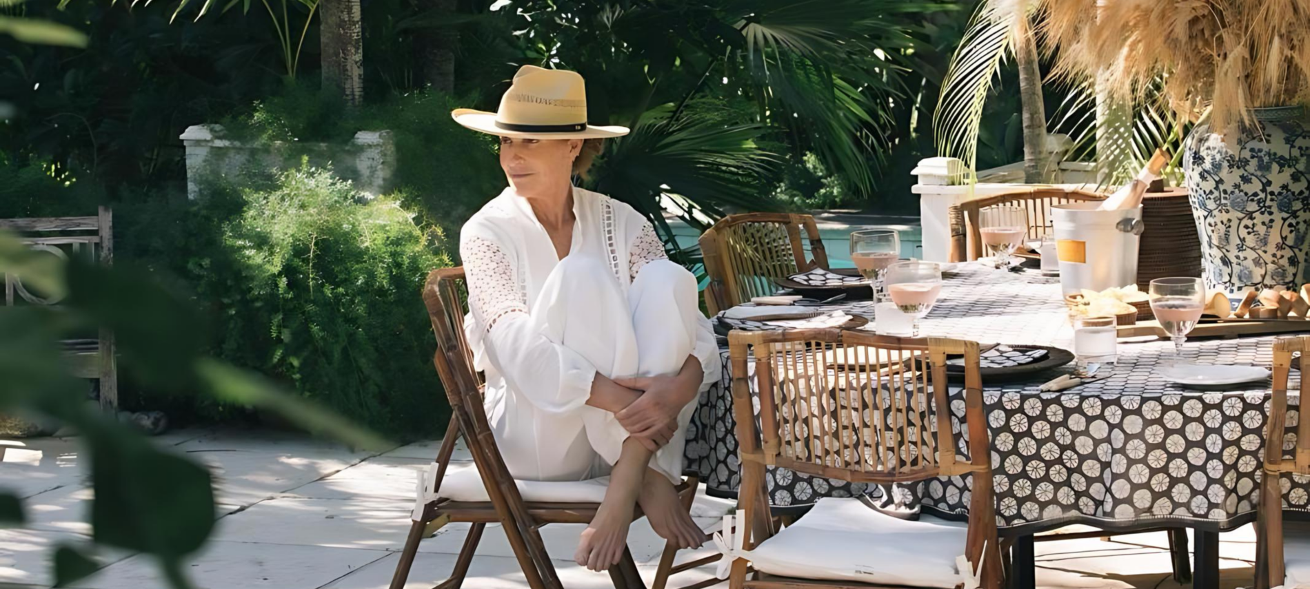 A serene alfresco dining scene with a stylish India Hicks in a white outfit and straw hat sitting thoughtfully at a beautifully set table amidst lush greenery