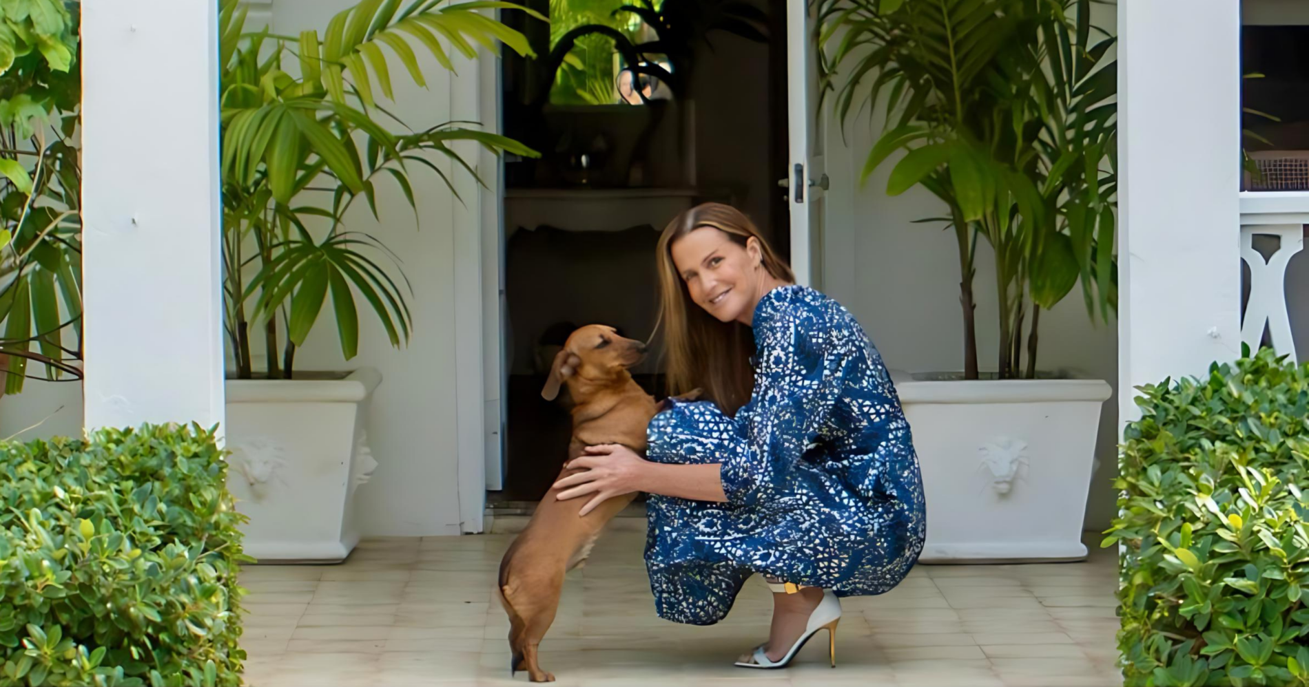 India Hicks in a blue patterned dress crouches down to lovingly embrace her brown dachshund in front of a white house entrance flanked by lush green plants.