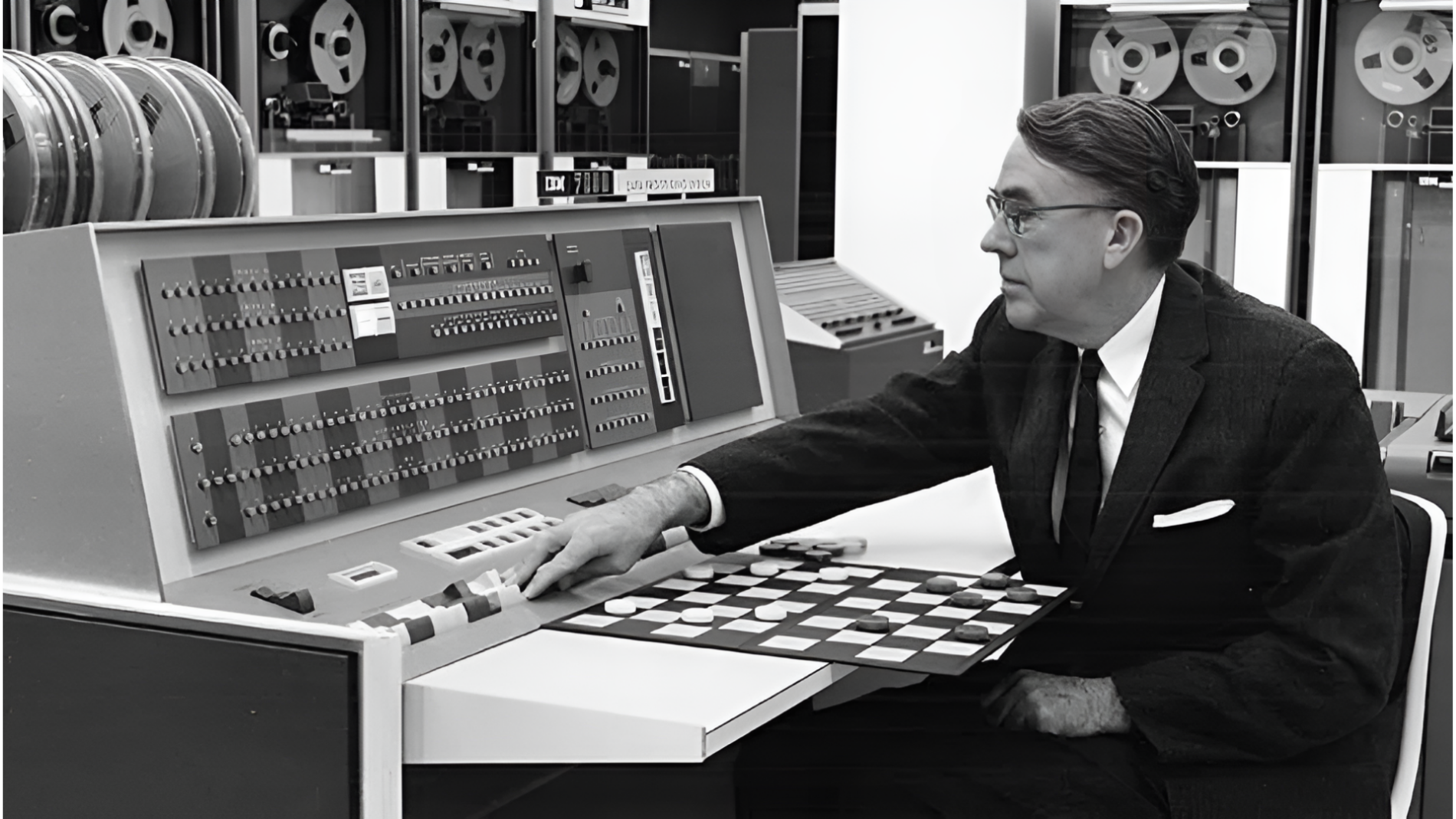 Arthur Samuel in a suit operating his Checkers programme onan early mainframe computer with reel-to-reel tape drives in the background.